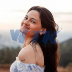 A young woman smiles warmly in an outdoor setting wearing a floral off-shoulder top.